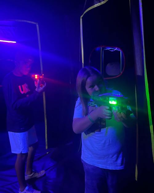 Two children in a dark room with neon lights, aiming laser tag guns. One has a red light, the other a green light.