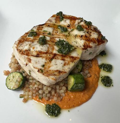 Grilled fish fillet on a bed of couscous and zucchini, topped with green sauce and served with a red sauce.