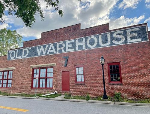 Brick building with large "OLD WAREHOUSE" sign, featuring red windows and a street lamp in front under a cloudy sky.