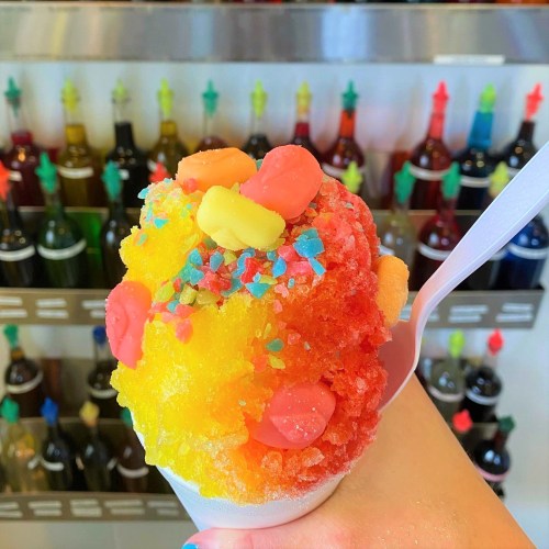 A colorful snow cone topped with candy and sprinkles, held in a hand against a backdrop of syrup bottles.