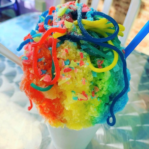 A colorful shaved ice treat topped with rainbow syrup and candy, served in a cup with two straws.