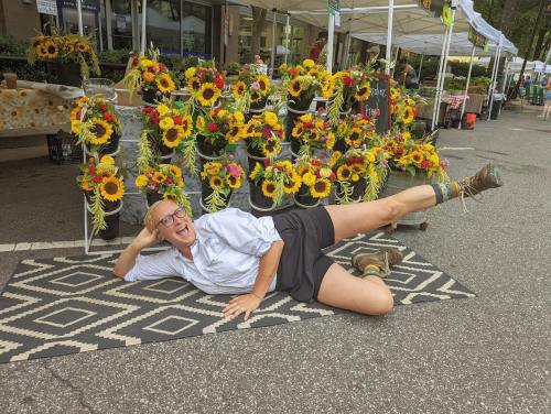 A person in a white shirt and shorts poses playfully on a rug beside vibrant sunflower arrangements at a market.