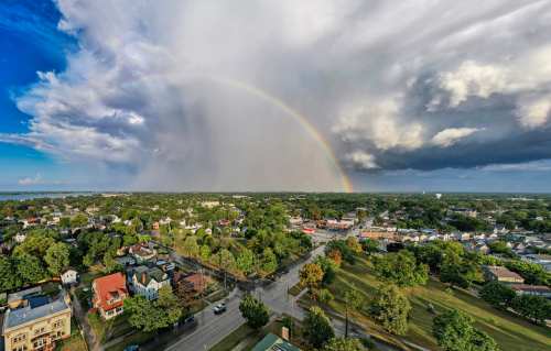 Aerial view of a town with a rainbow arching over dark clouds and greenery, showcasing a mix of sunlight and rain.