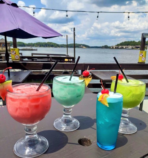 Four colorful cocktails with fruit garnishes sit on a table by a lake, under a cloudy sky.