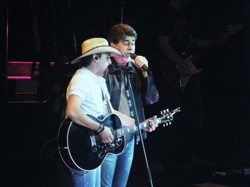 Two male musicians perform on stage, one with a guitar and a cowboy hat, the other singing into a microphone.