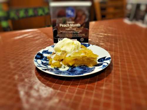 A plate of peach cobbler topped with vanilla ice cream, set on a blue floral plate with a sign about Peach Month in the background.