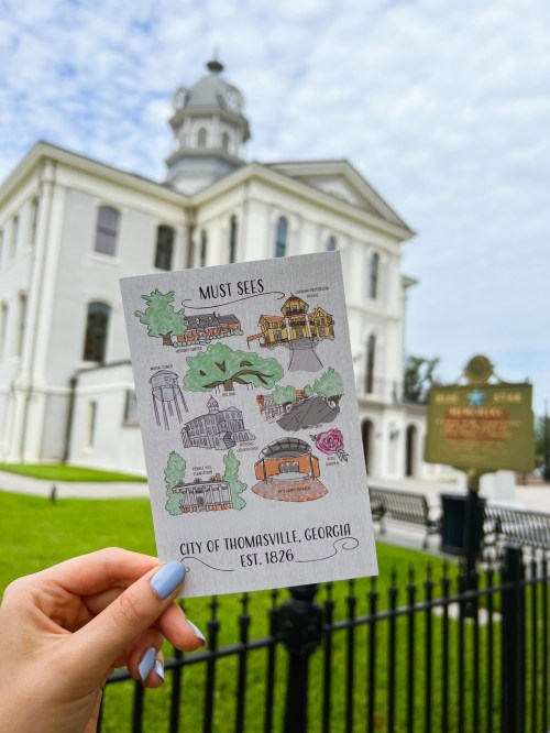 A hand holds a map of Thomasville, Georgia, with notable sites, in front of a historic white building.
