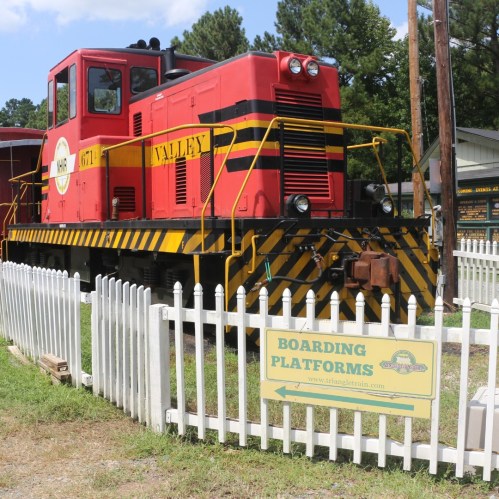 A red and yellow train engine parked beside a white picket fence with a sign for boarding platforms.