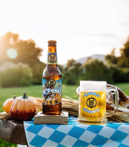 A bottle of DB beer and a glass of beer on a table with pumpkins and pretzels, set against a sunset backdrop.