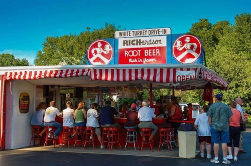 A busy drive-in restaurant with a red and white striped awning, featuring a sign for Richardson Root Beer and patrons at the counter.