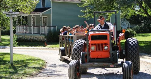 A tractor pulls a wagon full of people down a gravel road near a house on a sunny day.