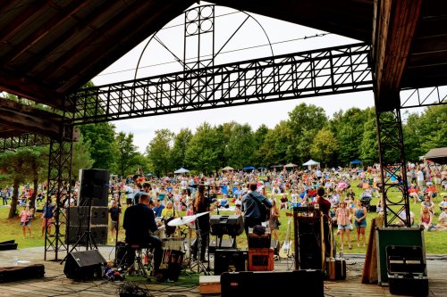 A band performs on stage in front of a large crowd at an outdoor music festival surrounded by trees.