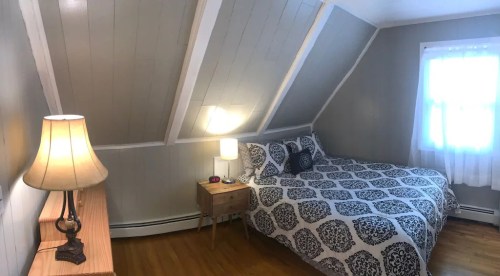 Cozy bedroom with sloped ceiling, a bed with patterned bedding, a nightstand, and a lamp, illuminated by natural light.