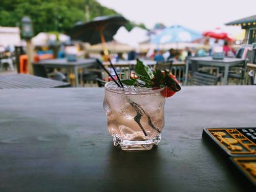 A refreshing cocktail with mint and a strawberry, served in a clear glass on a wooden table, with outdoor seating in the background.