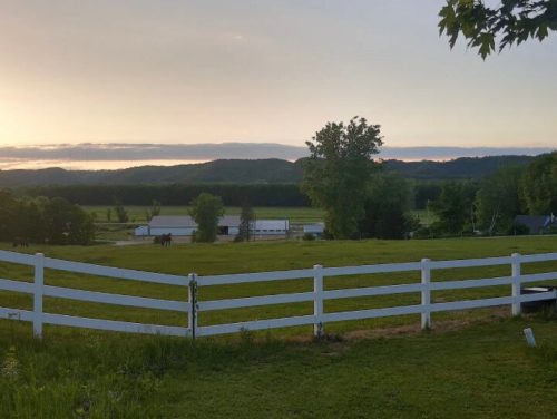 A serene landscape featuring a green field, white fence, and distant mountains under a sunset sky.