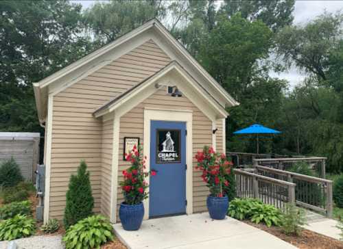 A small, charming chapel with blue doors, surrounded by greenery and flowers, featuring a deck and outdoor seating.