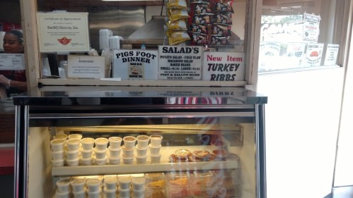 Counter display at a BBQ restaurant featuring salads, turkey ribs, and various side dishes in containers.