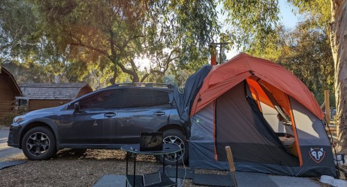 A car parked next to a tent in a campsite, surrounded by trees and sunlight filtering through.