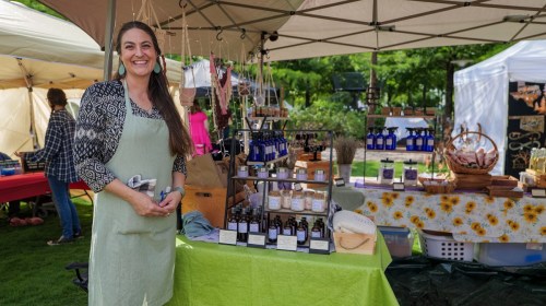 A smiling woman in an apron stands at a market stall filled with bottles and handmade products.