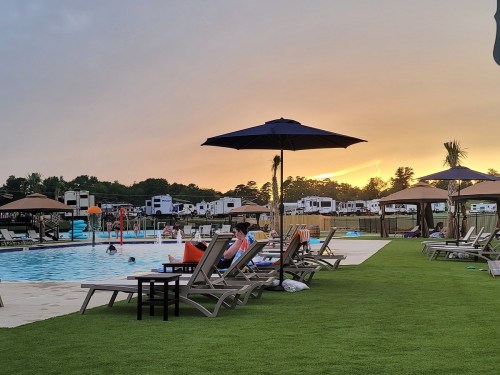 A serene pool area at sunset, featuring lounge chairs, umbrellas, and RVs in the background.