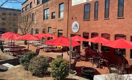Outdoor dining area with red umbrellas, brick walls, and tables in a sunny courtyard.