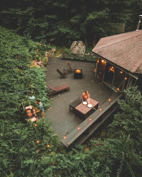 A cozy outdoor deck surrounded by greenery, with two people dining and string lights creating a warm atmosphere.