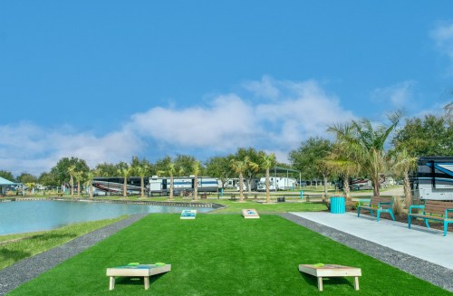 A grassy area with cornhole boards, surrounded by palm trees and RVs near a calm lake under a blue sky.