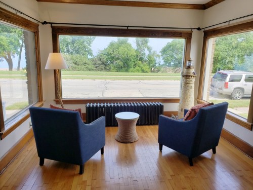 Cozy seating area with two blue chairs, a small table, and a view of trees and a street through large windows.
