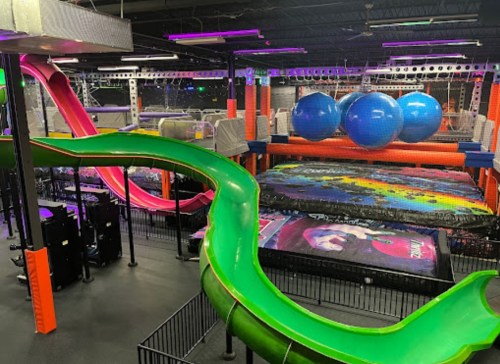 A colorful indoor trampoline park featuring green and pink slides, large blue balls, and various jumping areas.