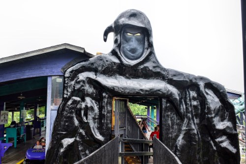 A large, dark figure resembling a grim reaper stands at the entrance of a ride, with a cloudy sky in the background.