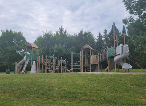 A playground with slides, climbing structures, and swings, surrounded by green grass and trees under a cloudy sky.