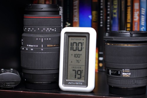 A digital thermometer displays indoor and outdoor temperatures next to two camera lenses on a shelf.