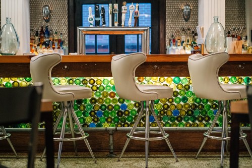 A stylish bar with three white stools, a colorful bottle display, and various beer taps in the background.