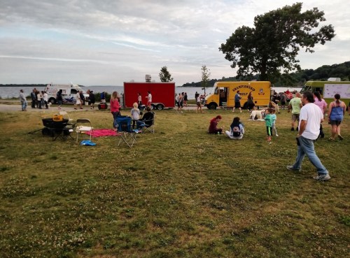 A lively outdoor scene by the water with food trucks, people mingling, and children playing on the grass.