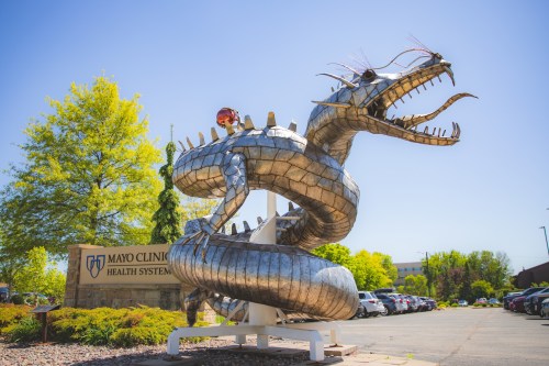 A large metallic dragon sculpture coils around a sign for Mayo Clinic Health System, set against a clear blue sky.