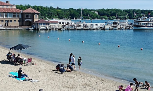 A sandy beach with people relaxing, walking, and a marina in the background on a sunny day.