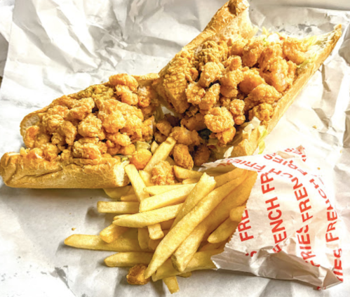 A large sandwich filled with fried shrimp, served with a side of golden French fries on crumpled paper.