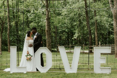 A couple in wedding attire embraces in front of large "LOVE" letters, surrounded by greenery and fairy lights.