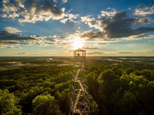 A silhouette of a person on a lookout tower against a sunset over a lush green landscape with clouds.