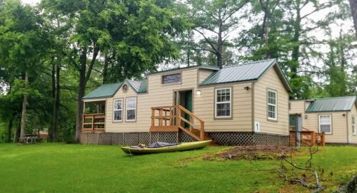 A cozy cabin with a green roof, surrounded by trees, and a kayak resting on the grass in front.