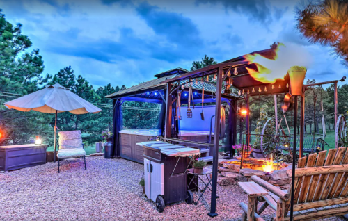 A cozy outdoor space with a gazebo, seating, grill, and fire pit, illuminated by soft lights at dusk.