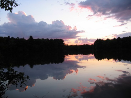 A serene lake at sunset, reflecting colorful clouds and trees in the calm water.