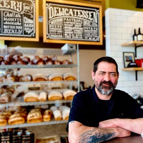 A smiling baker stands in front of a display of various breads in a cozy bakery setting.