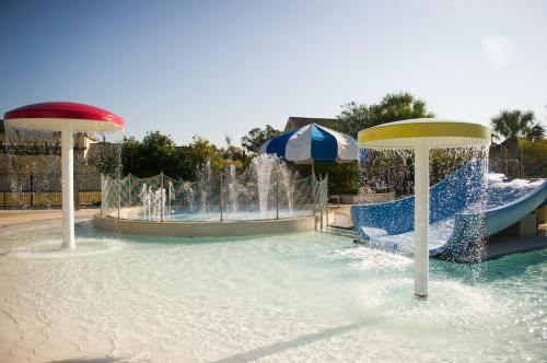 A sunny water park scene featuring colorful mushroom fountains and a water slide, surrounded by palm trees.