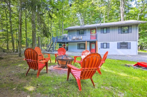 A cozy backyard with red chairs around a fire pit, surrounded by trees and a two-story house in the background.