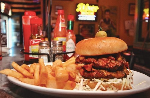 A stacked burger with crispy chicken, lettuce, and a pickle, served with golden fries on a plate in a diner setting.