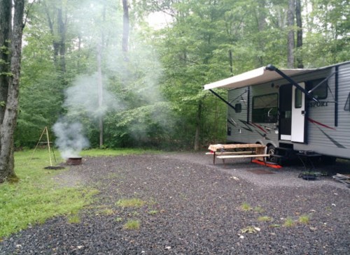 A camper parked in a wooded area with smoke rising from a fire pit nearby, surrounded by gravel and trees.