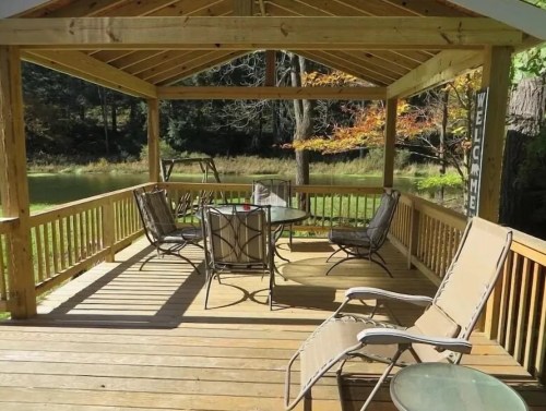 A wooden gazebo with a table and chairs, surrounded by greenery and trees, creating a welcoming outdoor space.