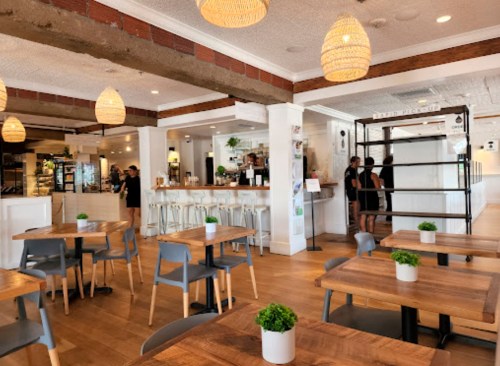 Bright, modern café interior with wooden tables, plants, and a bar area. Staff members are seen in the background.