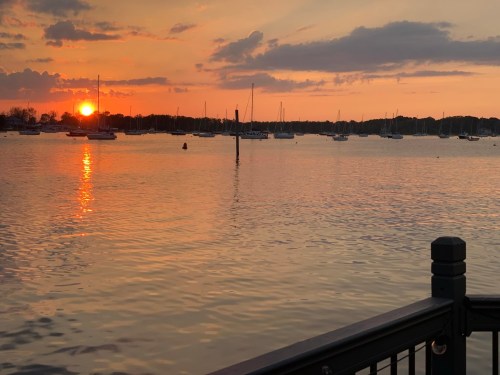 A serene sunset over a calm harbor, with boats anchored and reflections shimmering on the water.